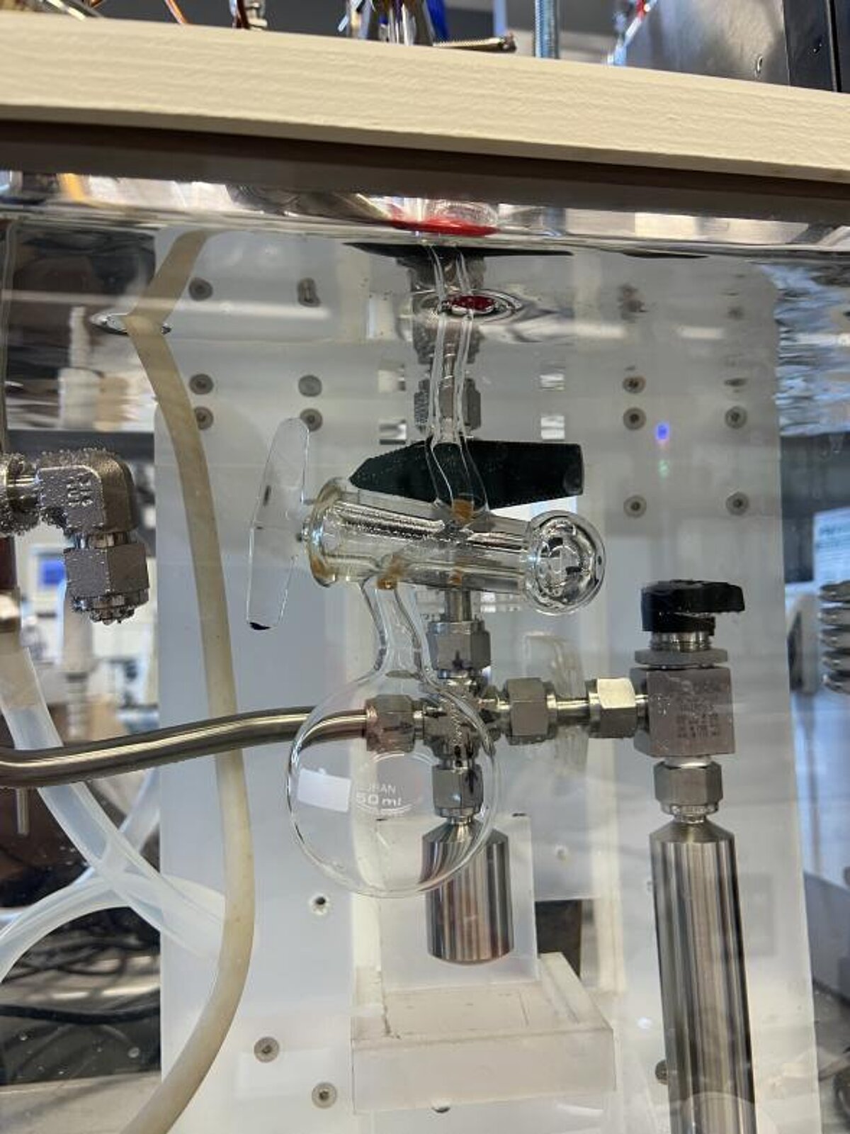 Detalizēts skats uz laboratorijas iekārtas fragmentu ar stikla traukiem, metāla savienojumiem un caurulēm; sistēma paredzēta gāzu šķīdības mērījumiem kontrolētos apstākļos.