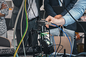 Laboratorijas vidē cilvēks zilā kreklā sakārto vadus uz sarežģīta zinātniska aparāta ar krāsainiem vadiem, ap viņu pulcējas skatītāji. Ierīce sastāv no zaļa lāzera, kas spīd uz lēcu un demonstrē lāzeru izmantošanu pētniecībā. 