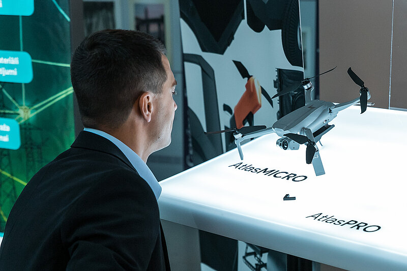 rsona skatās uz dronu, kas novietots uz balta stenda ar uzrakstiem 'AtlasMICRO' un 'AtlasPRO'. Fonā redzami informatīvi stendi ar grafiskiem elementiem