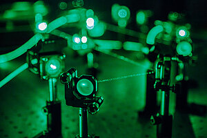 Zaļi lāzera staru kūļi šķērso tumšu laboratorijas telpu, apgaismojot dažādas optiskās lēcas un iekārtas. Šī aina rada fūturistisku, zinātnisku atmosfēru.