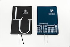 Divas piezīmju grāmatas uz balta fona: kreisā pusē ir melna ar lieliem burtiem "LU" un Latvijas Universitātes logo, pa labi ir zila ar ēkas ilustrāciju un LU logo