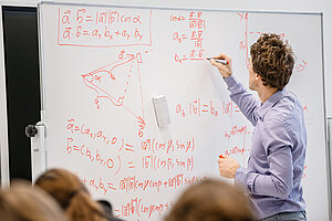 Cilvēks zilā kreklā, ar muguru pret kameru, raksta sarežģītas matemātiskas formulas uz tāfeles auditorijā. Auditorija ir pilna ar studentiem, kuri pieraksta pasniedzēja rakstītos vienādojumus. 