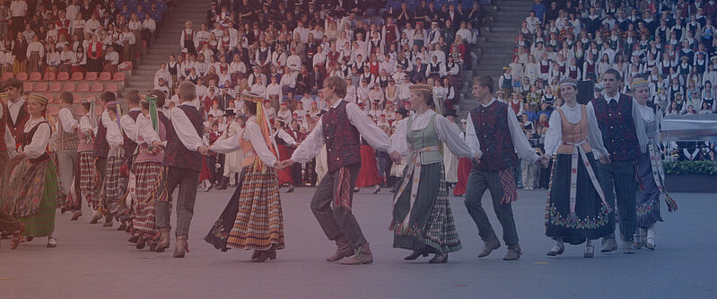 Dejotāju kopa tautastērpos dejo stadionā, turoties rokās, bet tribīnēs fonā sēž liels skatītāju un dalībnieku pulks