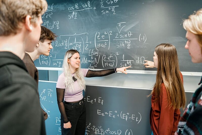 Skolēni stāv pie tāfeles ar matemātikas un fizikas vienādojumiem, diviem no viņiem aktīvi norādot uz formulām un skaidrojot risinājumu