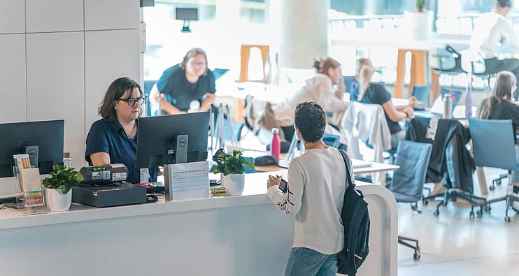 Cilvēks pie bibliotēkas informācijas letes runā ar darbinieku, fonā redzami galdi un lasītāji gaišā telpā.