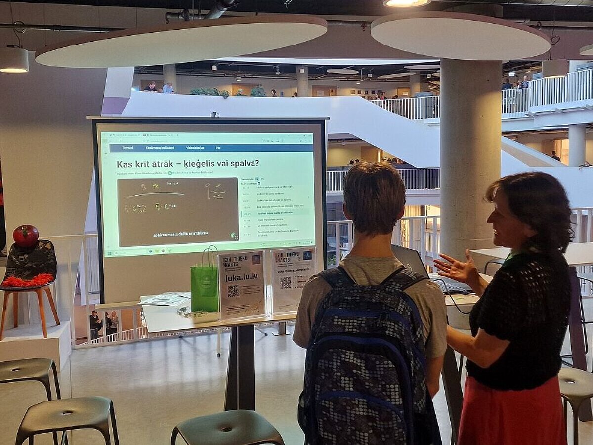 Skolēni novērtē luka.lu.lv mājaslapu, kurā apkopoti Kāna akadēmijas fizikas video ar transkriptiem un fizikas terminu skaidrojumiem latviešu valodā. Foto: Jeļena Kosmača, Zinātnieku nakts 2025 Latvijas Universitātē. Divi cilvēki stāv pie galda un skatās uz lielu projekcijas ekrānu, kurā redzams mācību materiāls ar virsrakstu 'Kas krīt ātrāk — ķieģelis vai spalva?'. Apkārt redzama moderna, gaiša telpa ar balkoniem un cilvēkiem fonā