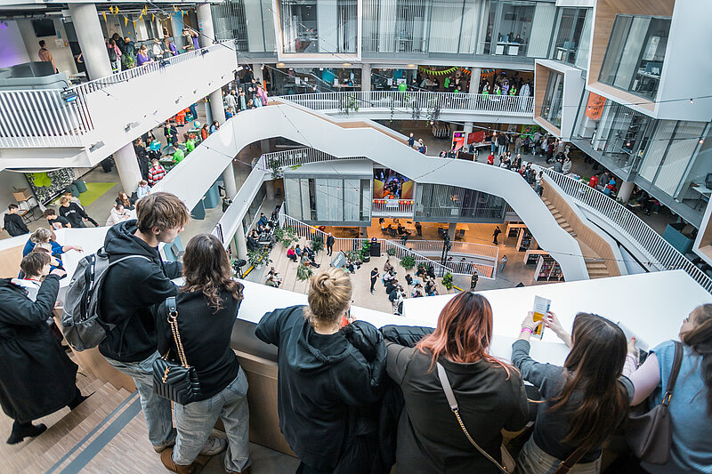 Vairāki jaunieši stāv augšējā stāvā LU Zinātņu mājā un noraugās uz norisēm zemāk