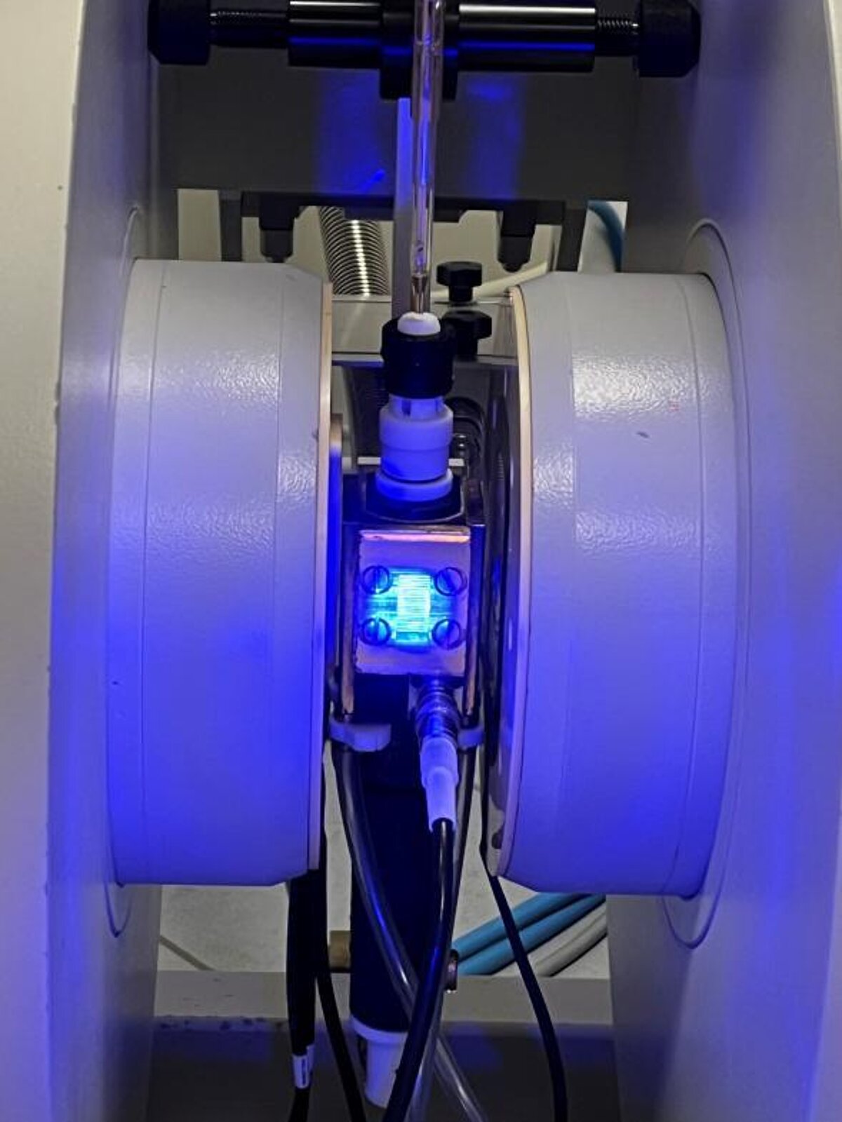 Zinātniska iekārta ar cilindriskām komponentēm un caurulēm; centrā redzams apgaismots mērījumu mezgls zilā gaismā, kas tiek izmantots jonu šķidrumu īpašību analīzei.