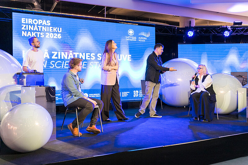 Diskusija uz Eiropas Zinātnieku nakts skatuves, kur vairāki dalībnieki sarunājas auditorijas priekšā.