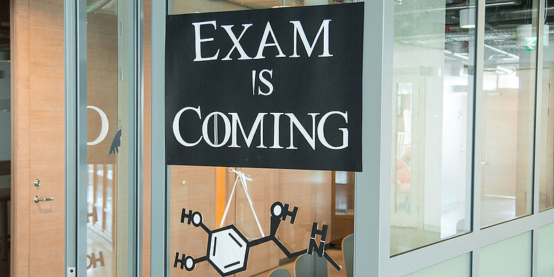 A poster on a glass wall with the phrase 'EXAM IS COMING' styled like the Game of Thrones slogan, with a chemical structure illustration below