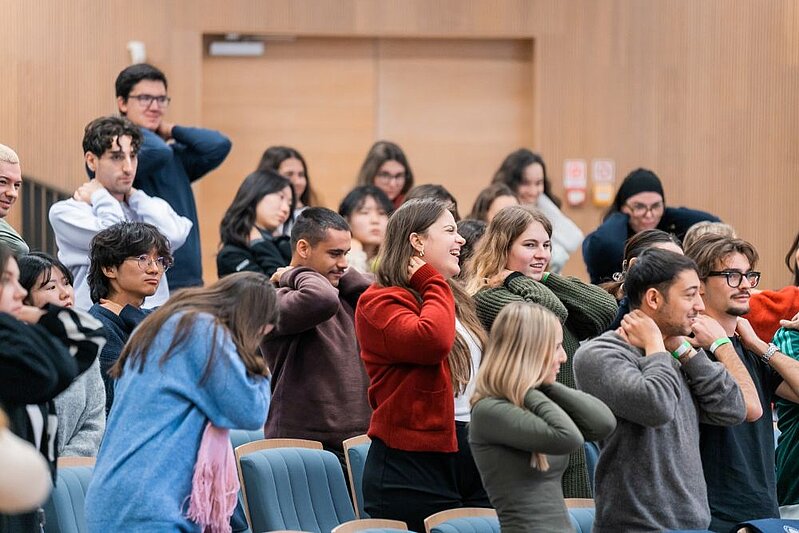 Studentu grupa auditorijā kopīgi piedalās vingrinājumā, turot rokas pie kakla vai pleciem, stāvot pie savām sēdvietām