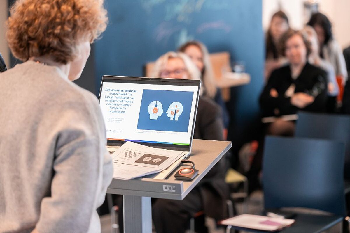 starptautiska konference “Doktorantu vadība pārmaiņu priekšā” – priekšplānā dators ar prezentāciju, fonā dalībnieki