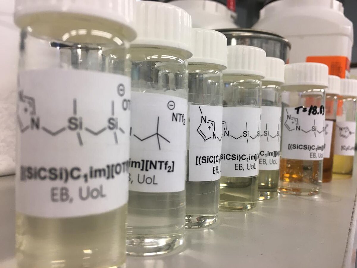 Stikla flakoni laboratorijas vidē, pildīti ar dažādas krāsas šķidriem ķīmiskiem savienojumiem; uz etiķetēm redzamas strukturālās formulas un paraugu apzīmējumi, flakoni novietoti rindā uz darba virsmas.