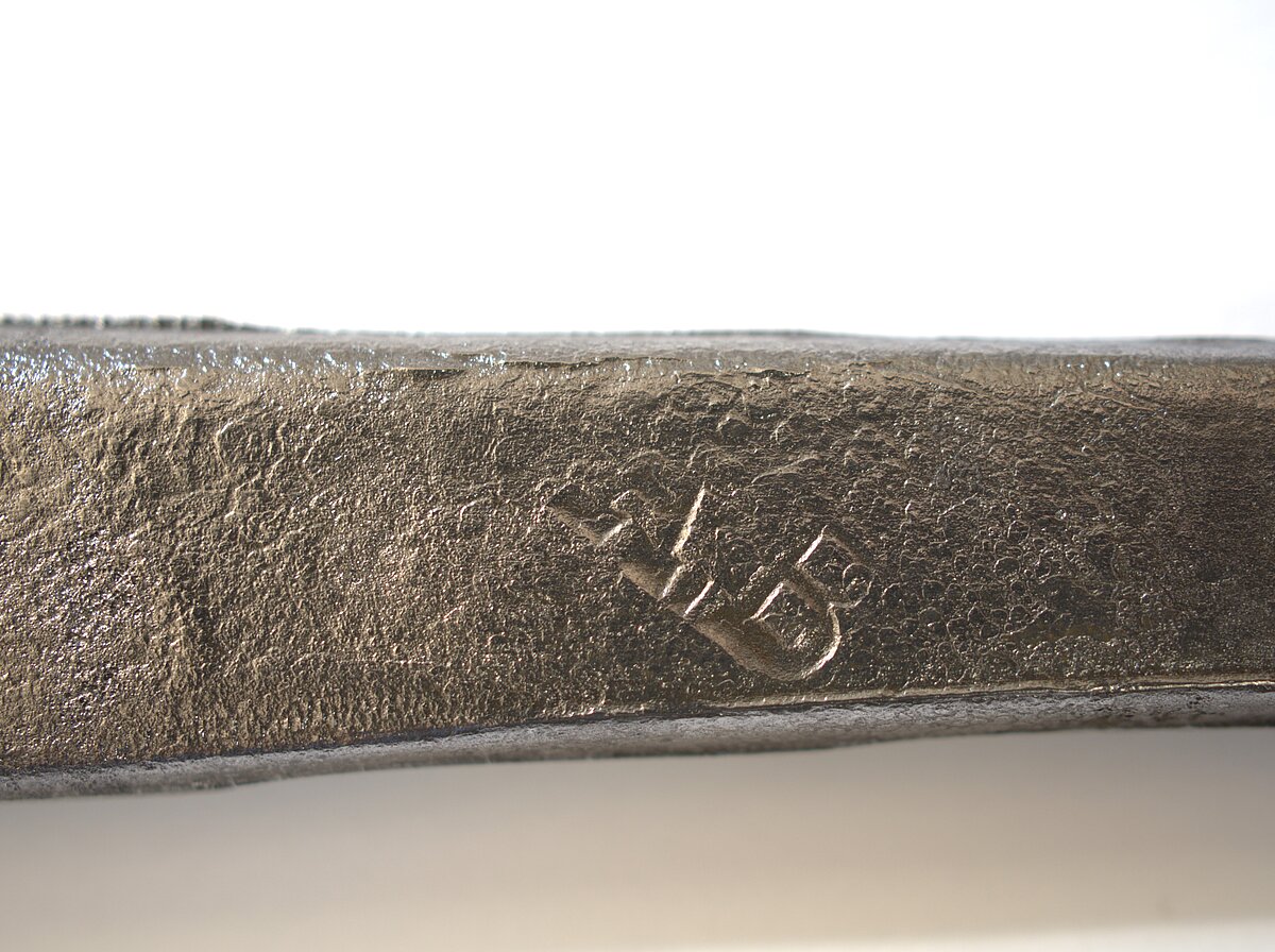 10. att. Notīrīts korozijas slānis un abreviatūra A.B.. uz rapiera asmens astes daļas. Foto: Gints Ajausks 10. att. Notīrīts korozijas slānis un abreviatūra A.B.. uz rapiera asmens astes daļas. Foto: Gints Ajausks