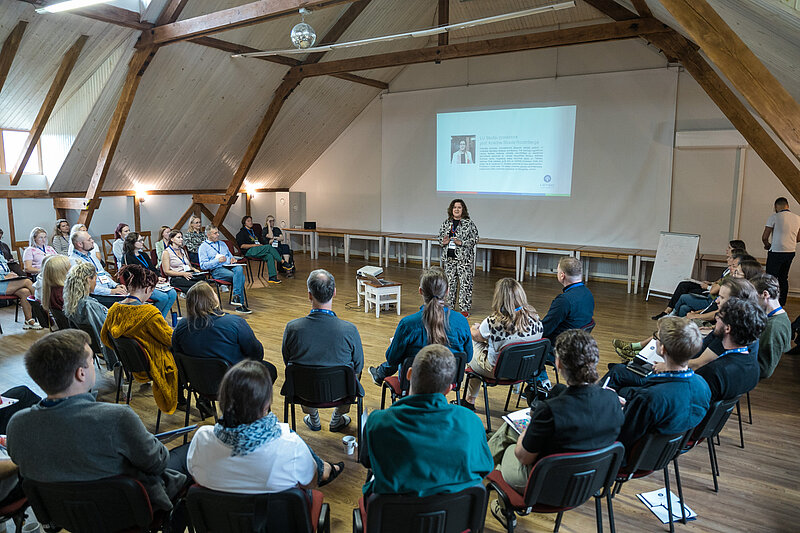 Cilvēku grupa sēž krēslos aplī koka siju telpā ar slīpu griestu konstrukciju; telpas priekšā stāv lektors un runā auditorijai, fonā redzams ekrāns ar prezentācijas slaidu, uz grīdas — projektors.