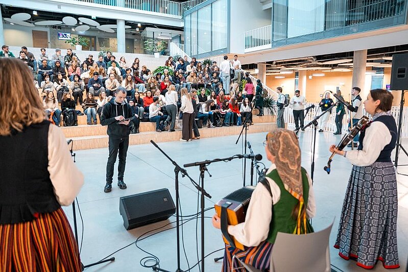 Plaša studentu auditorija pulcējusies universitātes atvērtajā ātrijā uz pasākumu, kam skatuves priekšpusē muzicē folkloras tērpos tērpti izpildītāji ar vijoli un akordeonu