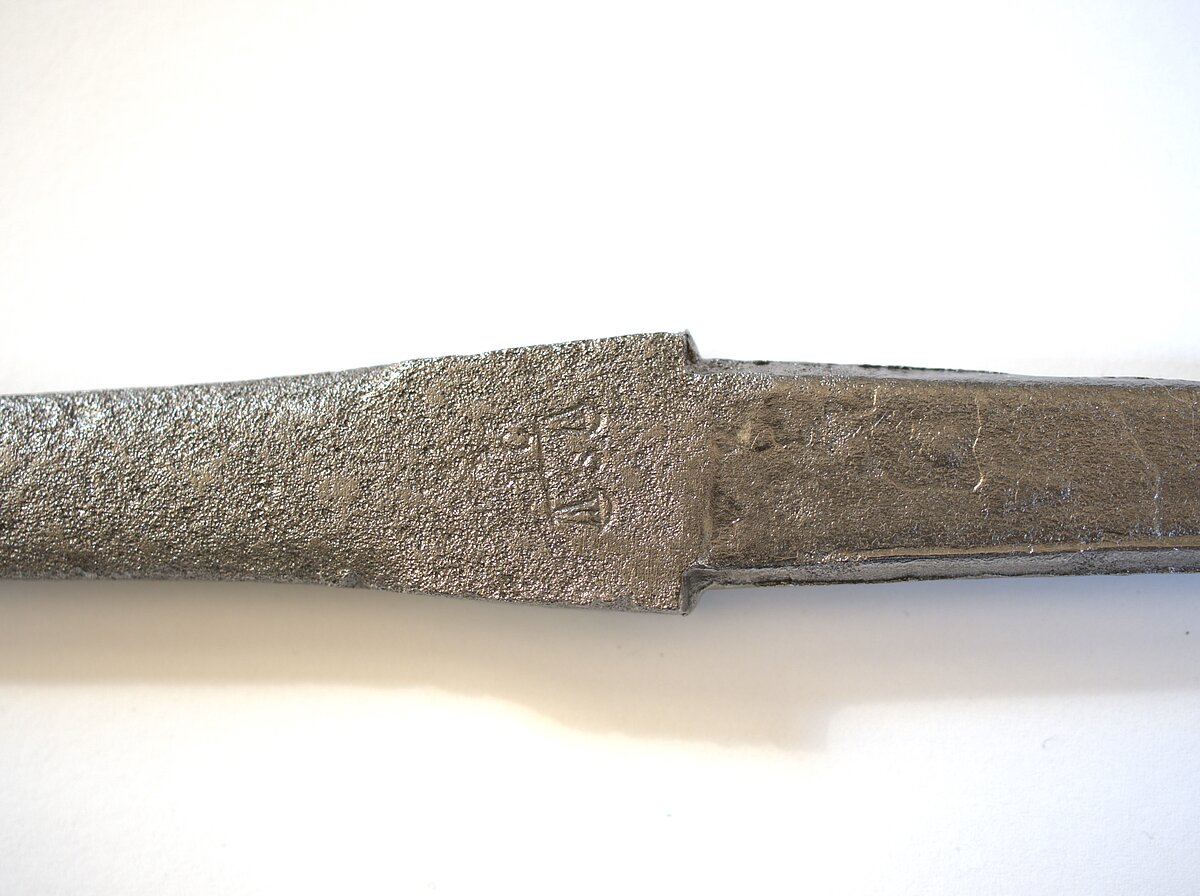 12. att. Notīrīts korozijas slānis un izgatavotāja vai meistara zīme uz rapiera asmens pleciem. Foto: Gints Ajausks 12. att. Notīrīts korozijas slānis un izgatavotāja vai meistara zīme uz rapiera asmens pleciem. Foto: Gints Ajausks