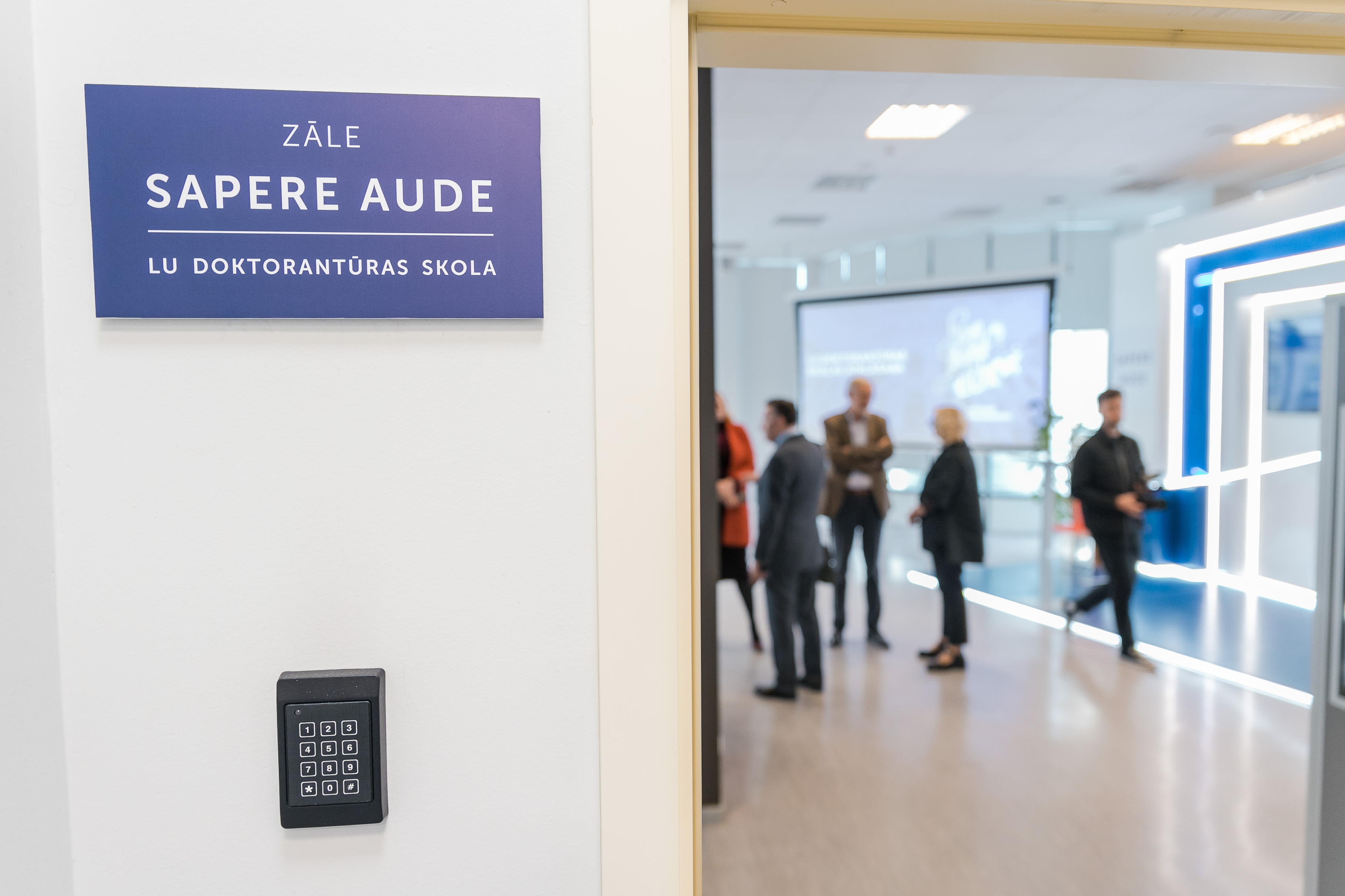 Plāksnīte pie sienas ar uzrakstu "Zāle Sapere Aude. LU Doktorantūras skola", fonā redzami cilvēki