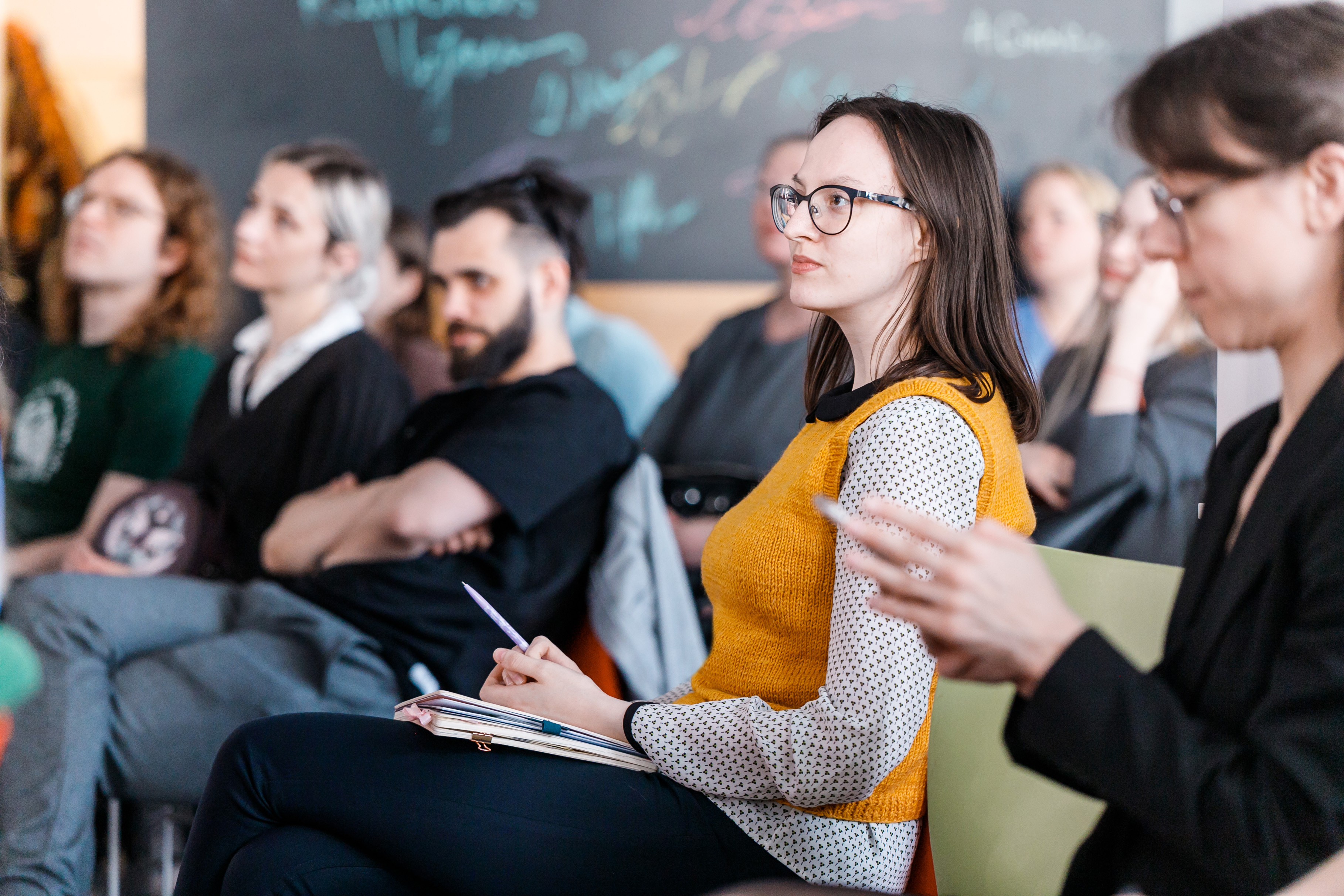 Jauna sieviete ar brillēm sēž auditorijā starp citiem studentiem