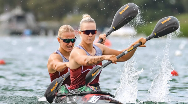Divas sportistes smaiļošanas divniekā ar airiem sacensību laikā uz ūdens, abām mugurā Latvijas izlases formas tērpi.