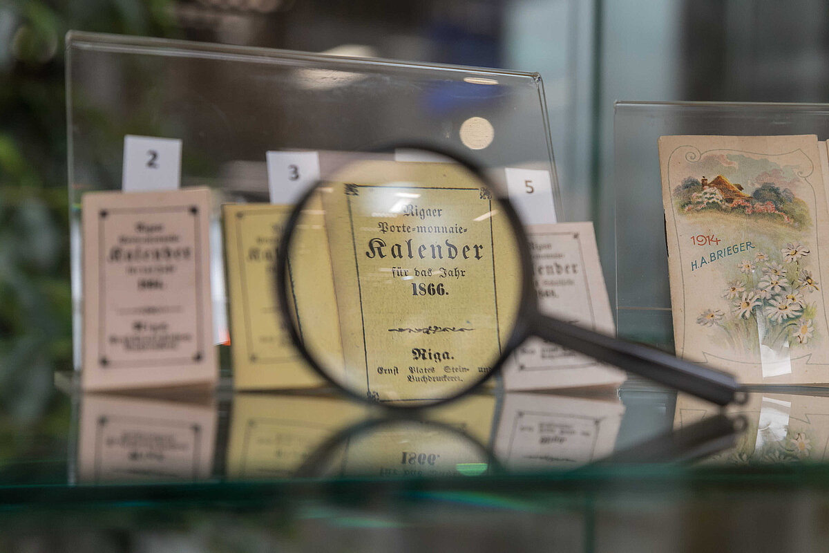 Stikla vitrīnā izstādīti vēsturiski kalendāri, no kuriem viens ar uzrakstu 'Rigaer Porte-monnaie Kalender für das Jahr 1866' redzams caur palielināmo stiklu. Fonā redzami citi nelieli kalendāri, tostarp viens ar gleznotu ainavu un uzrakstu '1914 H. A. Brieger