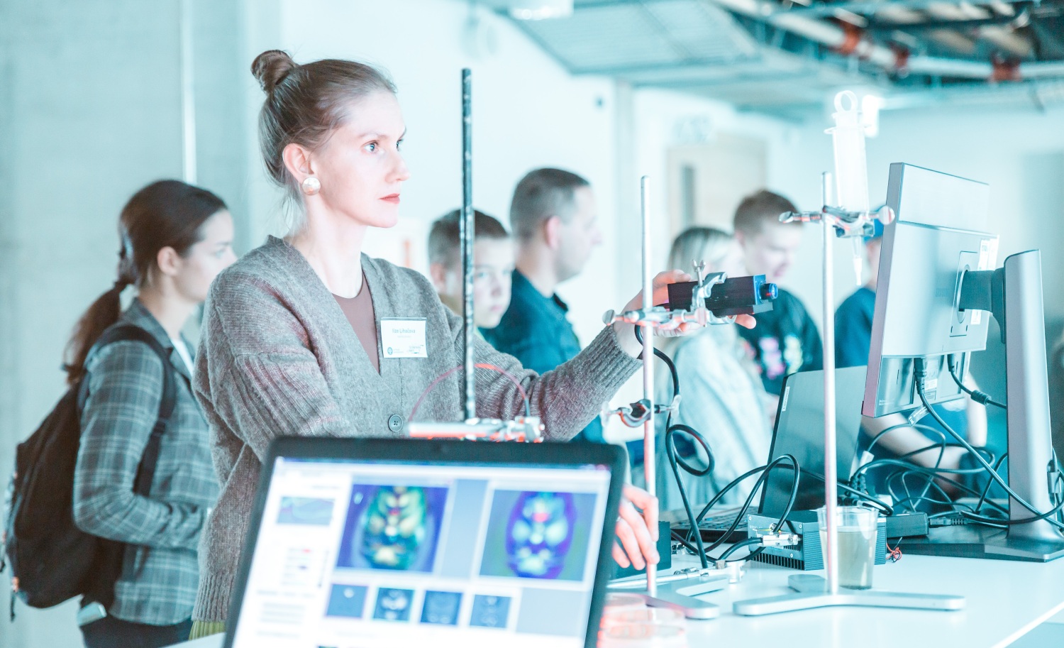 Pētnieki laboratorijā strādā ar zinātnisko aprīkojumu; priekšplānā redzams portatīvais dators ar smadzeņu skenēšanas attēliem, fonā kolēģi pie datoriem