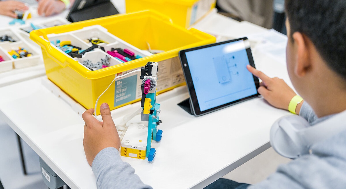Jaunietis sēž pie galda un konstruē robotu no LEGO detaļām, izmantojot planšetdatoru ar instrukcijām un dzeltenu LEGO komplekta kasti fonā