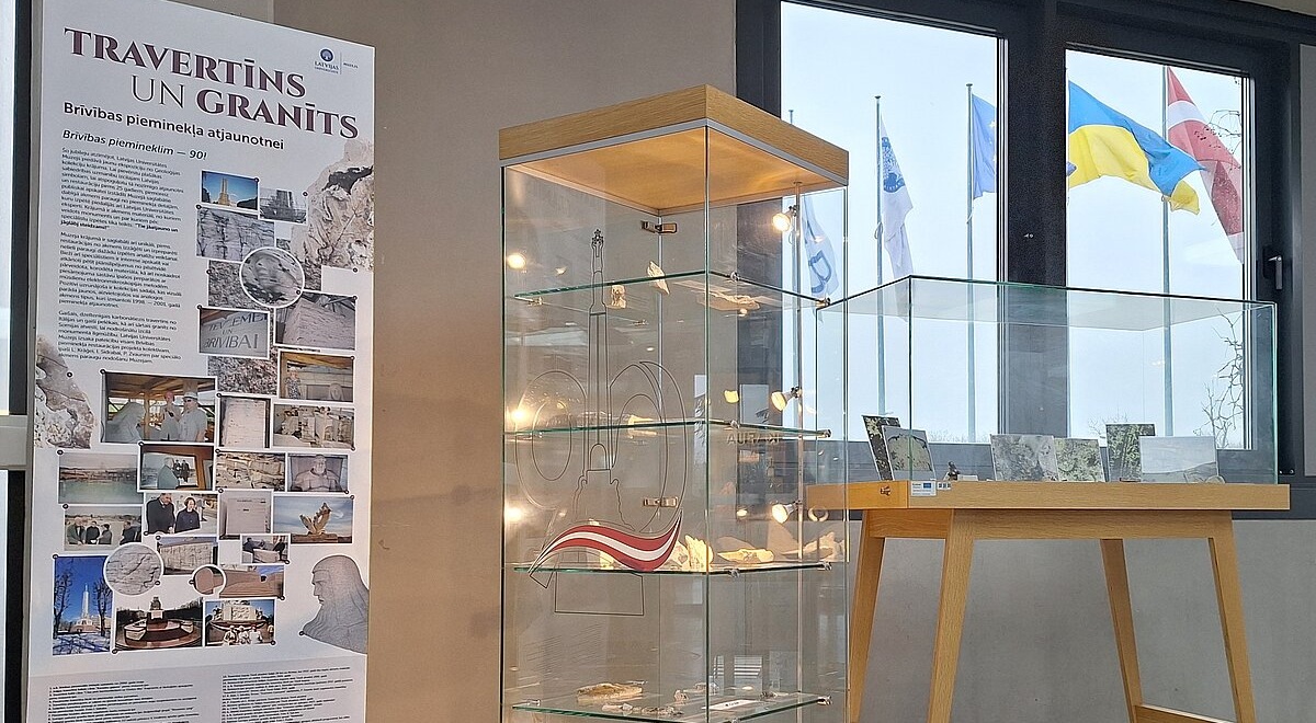 Izstādes stends ar informācijas plakātu “Travertīns un granīts. Brīvības pieminekļa akmeņi” kreisajā pusē un stikla vitrīnām ar akmens paraugiem un Latvijas karoga lentīti labajā pusē. Fonā redzami logi ar karogiem ārpusē.