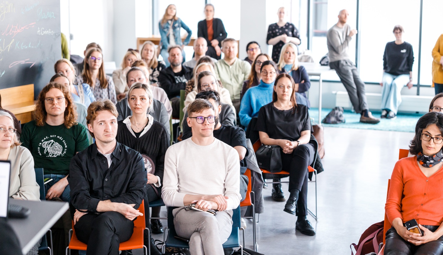 Cilvēku grupa sēž un stāv gaišā telpā, klausoties pasākuma prezentācijā