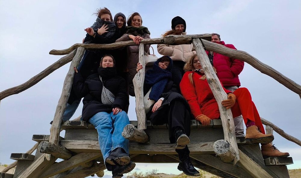 Cilvēku grupa pozē uz koka skatu platformas pie dabas takas, sēžot un stāvot uz konstrukcijas dažādos līmeņos ziemīgā, mākoņainā dienā