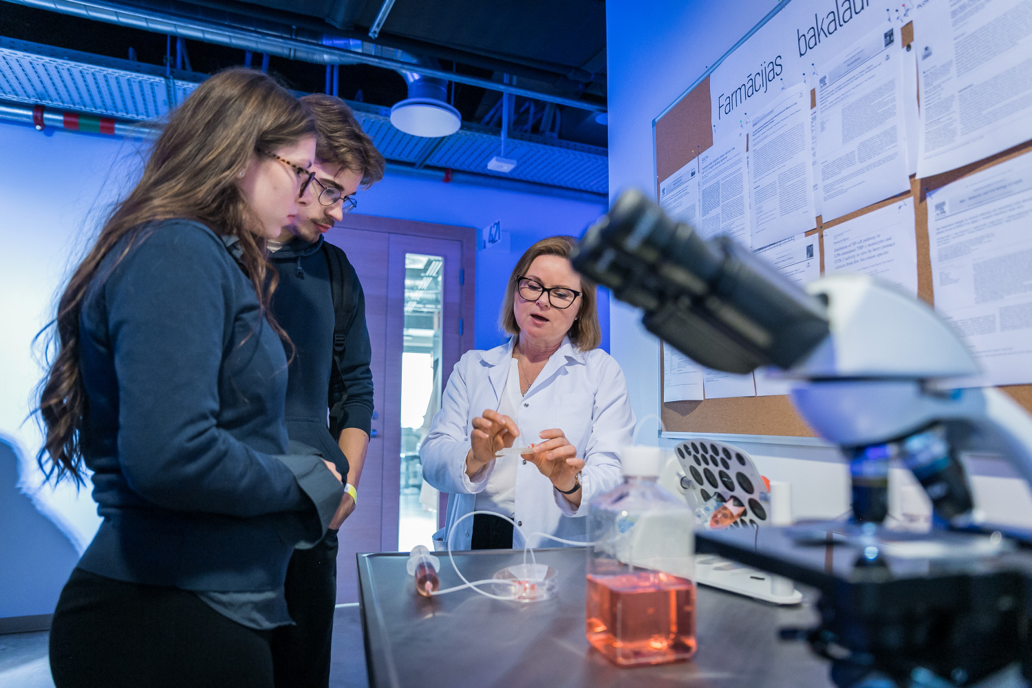 Laboratorijas telpa ar mikroskopu priekšplānā un zinātniskiem materiāliem uz galda. Fonā persona baltā halātā demonstrē eksperimenta aprīkojumu diviem interesentiem