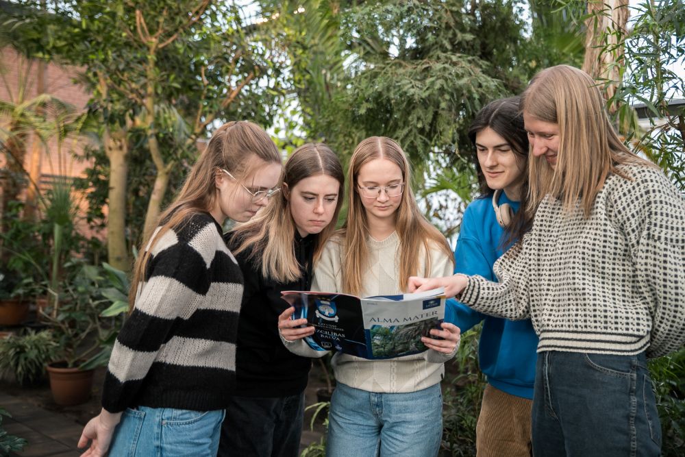 Pieci jaunieši stāv Botāniskajā dārzā starp augiem un kopīgi aplūko atvērtu Alma Mater žurnālu, ko tur viens no viņiem