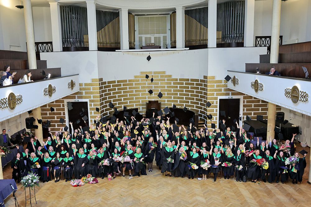 Liela absolventu grupa akadēmiskajos tērpos ar zaļiem šallēm pozē svinīgajā zālē, daudzi paceļot gaisā cepures un turot ziedus. Fonā redzama plaša skatuve un balkoni ar skatītājiem