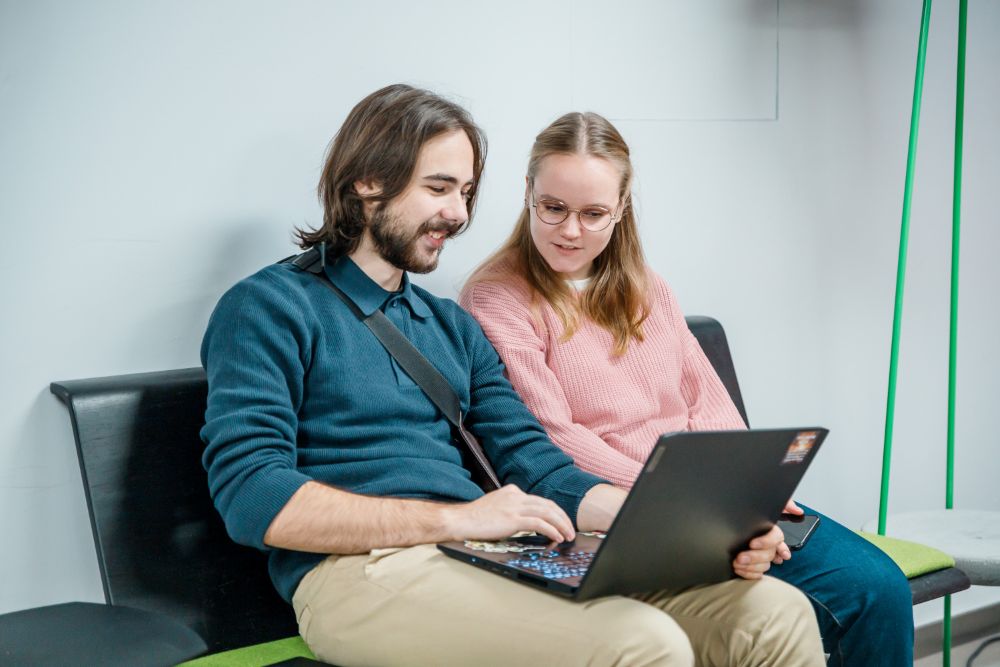 Divi cilvēki sēž uz soliņa pie sienas un kopīgi skatās portatīvajā datorā