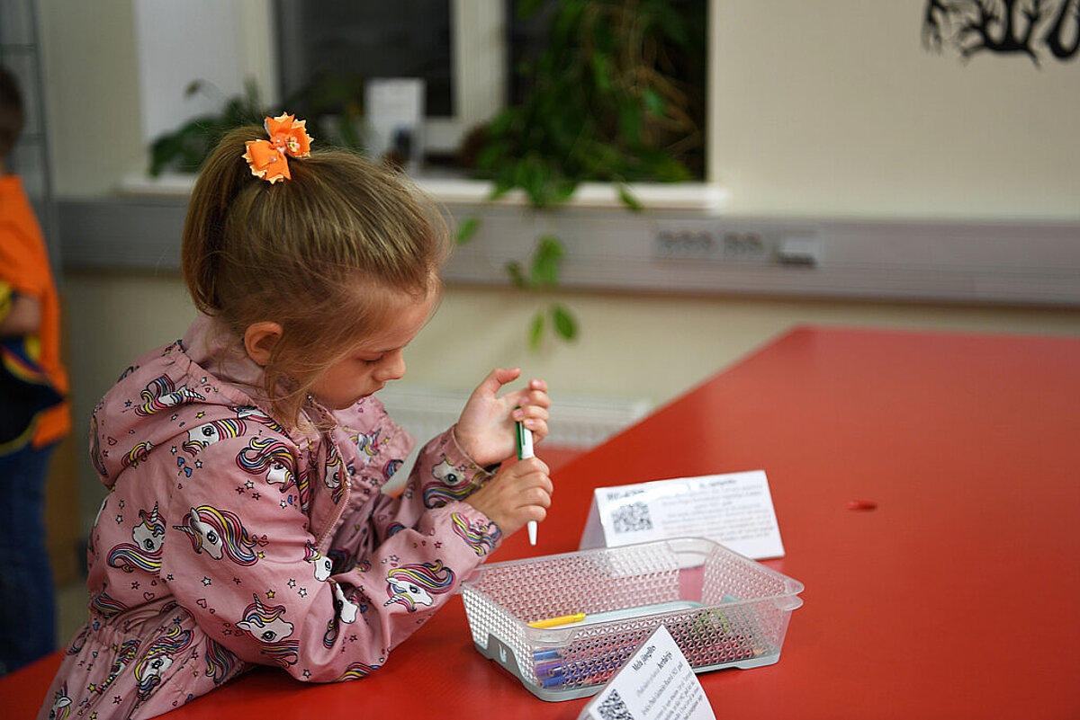Bērns rozā jakā ar vienradžu rakstu sēž pie sarkana galda un tur rokās marķieri. Priekšā atrodas caurspīdīga kaste ar krāsainiem zīmuļiem un citiem materiāliem, kā arī informatīvas kartītes