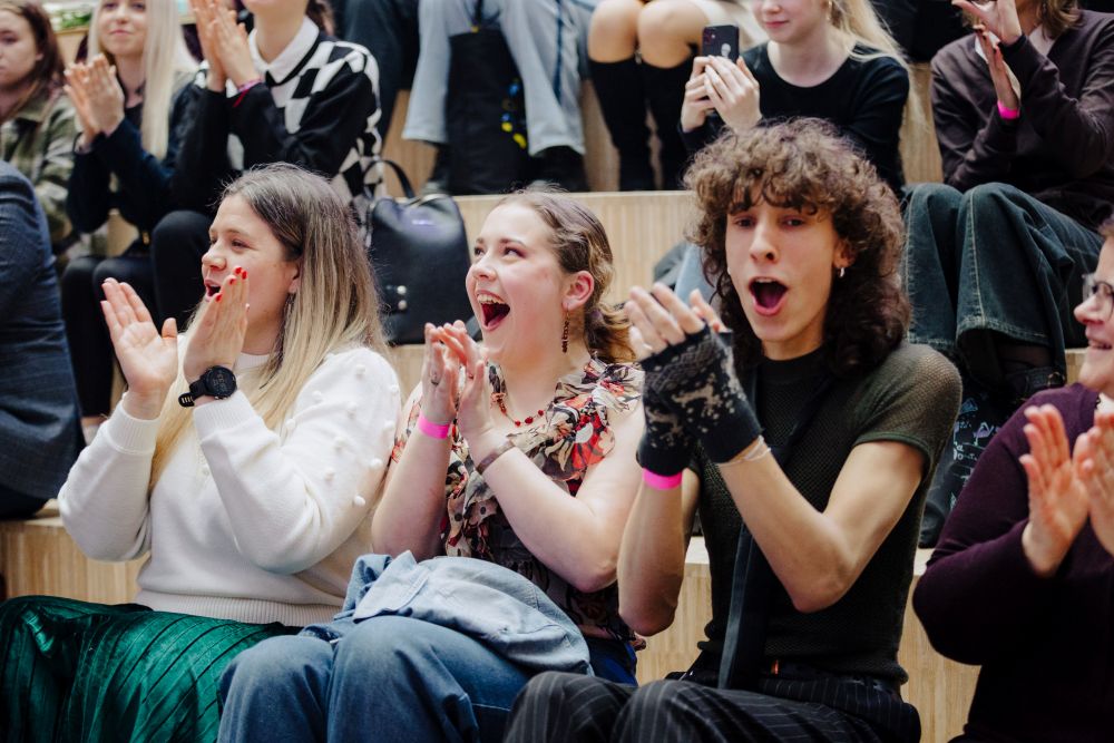Pasākuma apmeklētāji sēž uz pakāpienu tipa sēdvietām un aktīvi aplaudē, radot dzīvīgu un atbalstošu atmosfēru.