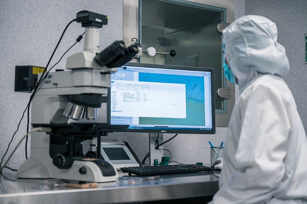 Laboratorijas darbinieks aizsargtērpā strādā pie datora ekrāna, blakus atrodas mikroskops un cits laboratorijas aprīkojums.