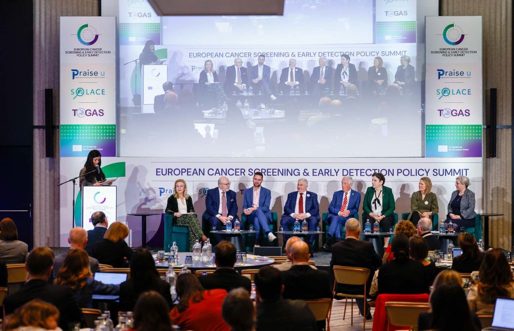 Konferences zālē notiek Eiropas vēža skrīninga un agrīnas atklāšanas politikas samits. Uz skatuves sēž deviņi paneļdiskusijas dalībnieki, savukārt kreisajā pusē pie tribīnes runā moderatore. Aiz viņiem redzams liels ekrāns ar paneļdiskusijas tiešraides attēlu. Auditorija sēž zālē un klausās