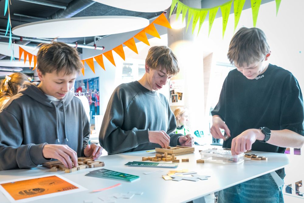 Jaunieši stāv pie galda un darbojas ar koka elementiem un spēļu detaļām radošā aktivitāšu zonā, kur telpu rotā košas karodziņu virtenes.