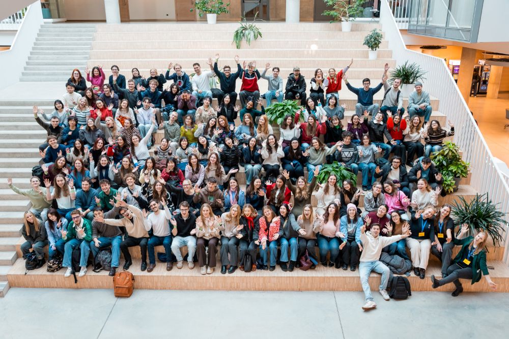 Liela studentu un pasniedzēju grupa sēž un stāv uz plašām, gaišām kāpnēm modernā universitātes ēkā, pozējot kopbildei un pacilāti mājot kamerai.