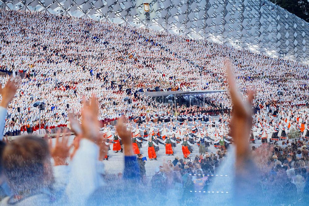 Liels Dziesmu un deju svētku skatuves laukums, kur tūkstošiem dalībnieku baltos tērpos dzied tribīnēs, bet priekšplānā dejotāji tautas tērpos veido rakstainas rindas; skatītāju rokas redzamas kadrā priekšā