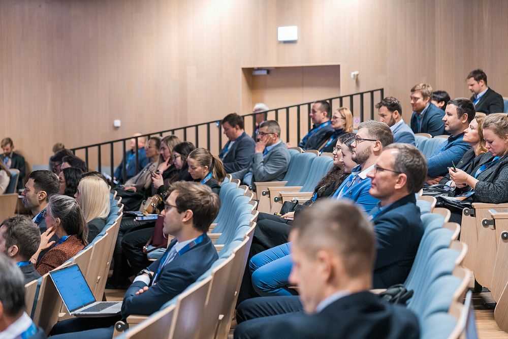 Plaša auditorija ar cilvēkiem, kas sēž rindās un klausās pasākumu vai prezentāciju. Telpā ir koka paneļu sienas un vairākas sēdvietu rindas
