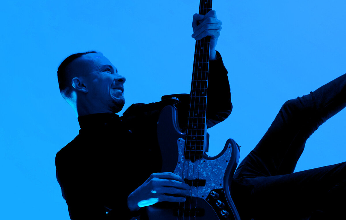 Persona tumšā apģērbā dinamiskā pozā spēlē elektrisko basģitāru uz spilgti zilā fona.