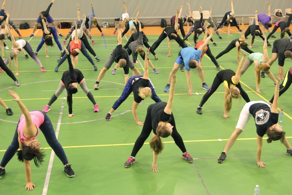 Sporta zālē liela cilvēku grupa veic iesildīšanās vingrinājumu, stāvot plati kājās un noliecoties uz sāniem ar vienu roku izstieptu augšup. Dalībnieki izvietoti vairākās rindās uz zaļa seguma laukuma