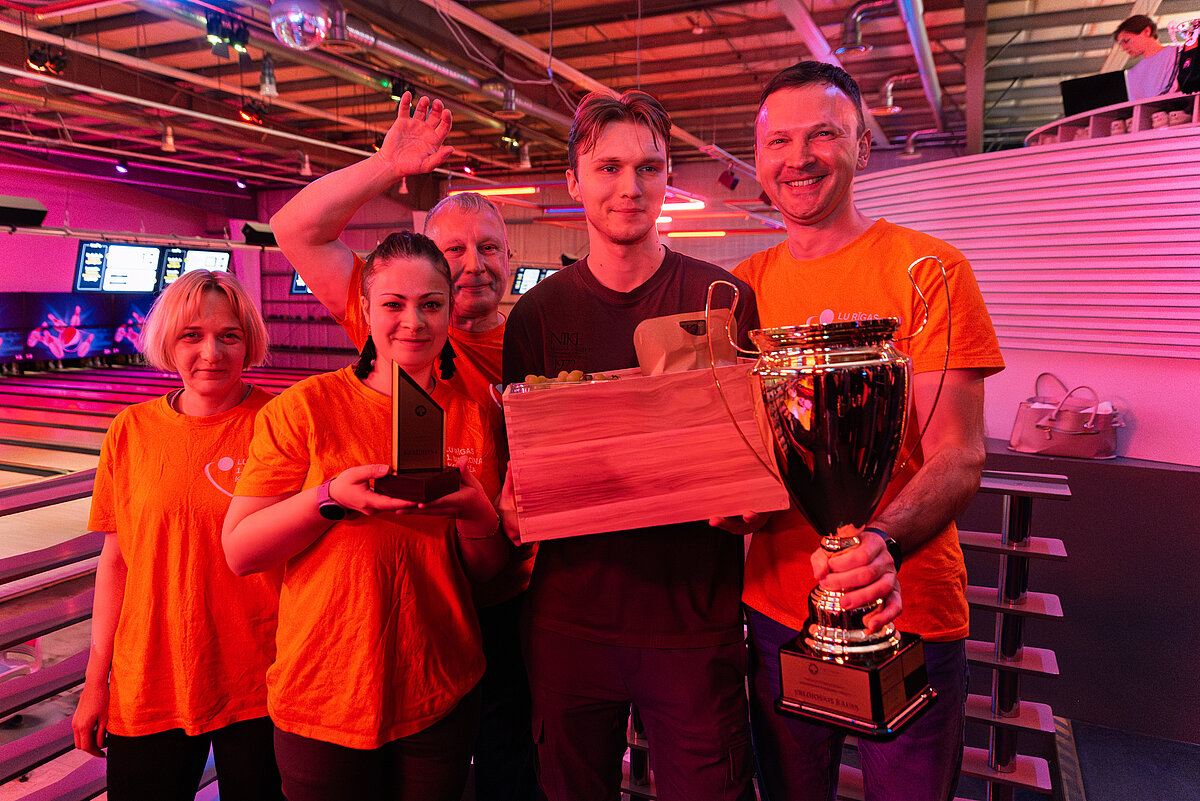 Cilvēku grupa boulinga zālē stāv kopā pie celiņiem, turot rokās lielu trofeju un dāvanu kasti. Daži dalībnieki valkā koši oranžus krekliņus. Fonā redzami boulinga celiņi, bumbas un ekrāni ar rezultātiem.