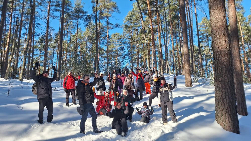 Liela cilvēku grupa ziemas apstākļos stāv un sēž sniegā priežu mežā. Visi ir silti ģērbušies un ar paceltām rokām pozē kamerai. Apkārt ir biezs sniegs, saules gaisma spoži izgaismo kokus un cilvēkus.