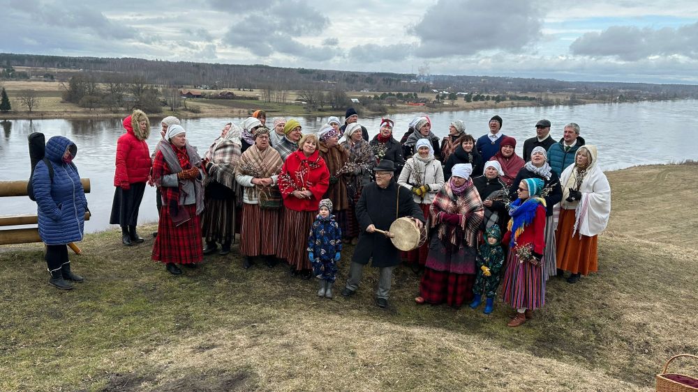 Liela cilvēku grupa tautas tērpos un ziemas apģērbā stāv uz nogāzes pie upes. Daļa dalībnieku tur tradicionālos mūzikas instrumentus un pavasara rotājumus. Fonā redzama plaša ainava ar upi, laukiem un mākoņainām debesīm