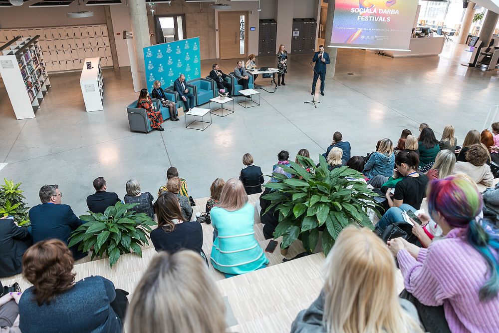 No auditorijas augšējām sēdvietām skatāms pasākums plašā, gaišā telpā. Priekšā uz skatuves zonas atrodas vairākas mēbeles un sēž vairāki paneļdiskusijas dalībnieki, bet viens runātājs stāv pie mikrofona. Auditorijā sanākušie klausītāji sēž pakāpienveida tribīnēs. Telpā redzami augi, bibliotēkas plaukti un ekrāns ar uzrakstu ‘Sociālā darba festivāls’