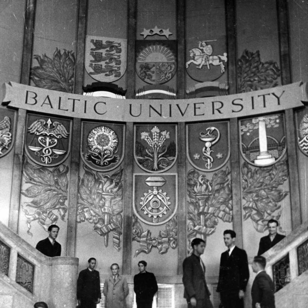 Vēsturiska fotogrāfija ar cilvēkiem, kas stāv plaša iekštelpu kāpņu telpā zem lielas dekoratīvas sienas ar Baltijas Universitātes nosaukumu un vairākiem vairogiem un emblēmām. Dekori attēlo dažādus Baltijas reģiona simbolus un fakultāšu zīmes