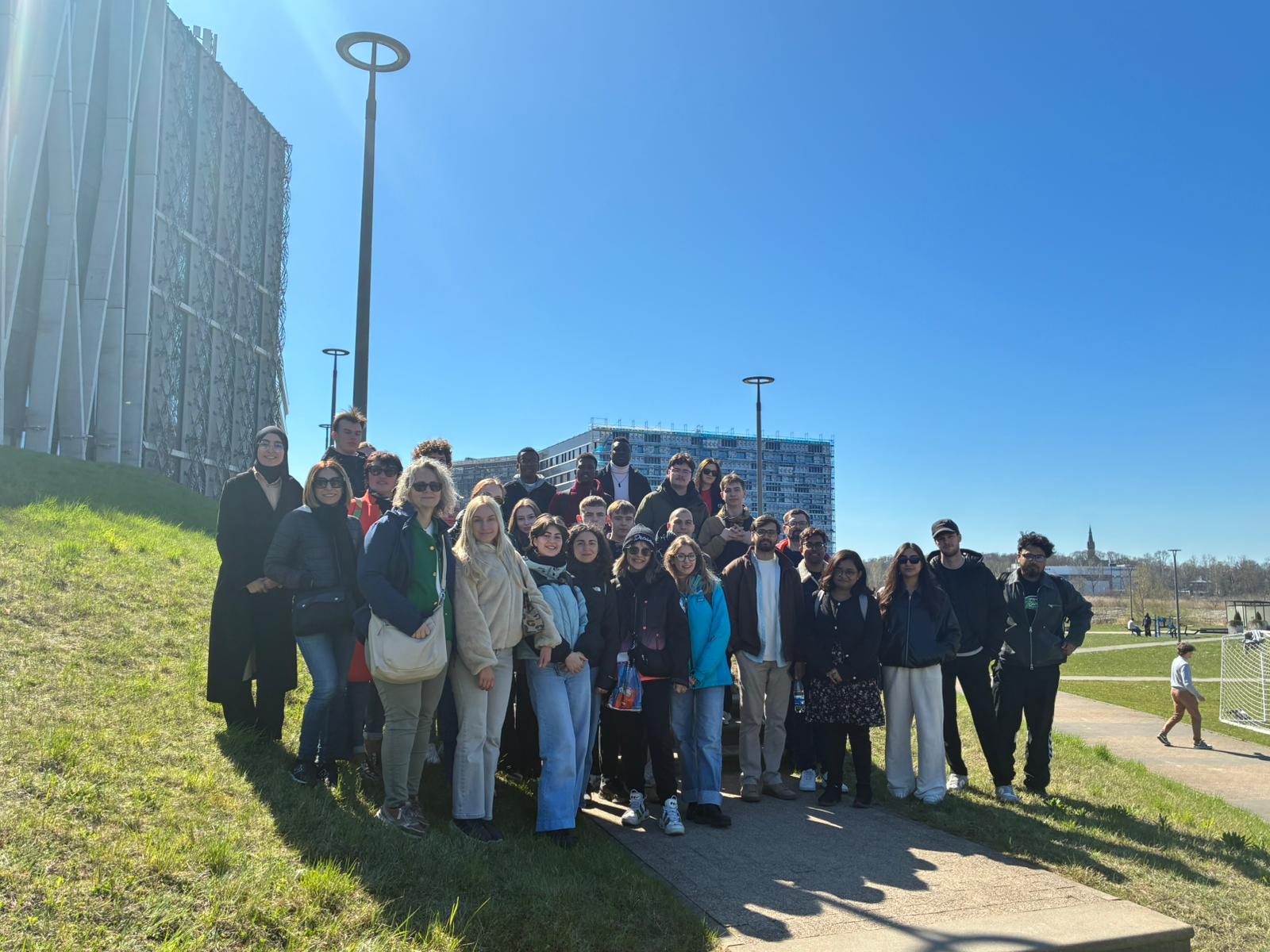 Cilvēku grupa stāv uz zāliena pilsētvidē saulainā laikā, fonā mūsdienīga biroju ēka un gājēju celiņi.